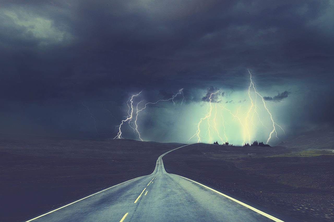Eine gerade Straße führt durch eine düstere Landschaft mit Gewitter und Blitzen im Hintergrund.