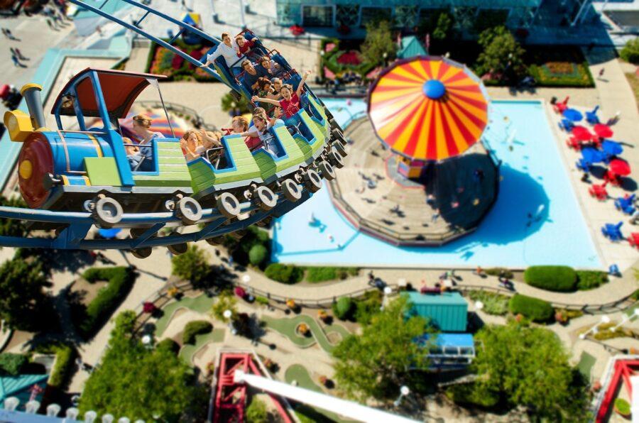 Eine Achterbahn mit Fahrgästen, die über einen Freizeitpark mit einem bunten Pavillon und einem Pool fährt.