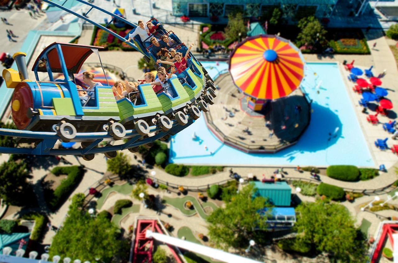 Eine Achterbahn mit Fahrgästen, die über einen Freizeitpark mit einem bunten Pavillon und einem Pool fährt.
